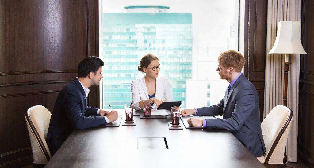 Portrait of young female business leader sitting in modern office and having meeting with her team. She showing something on tablet computer and asking questions